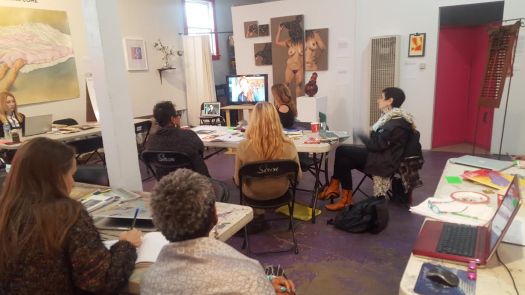 An art gallery with paintings on the walls, groups of women with laptops facing a large TV screen 