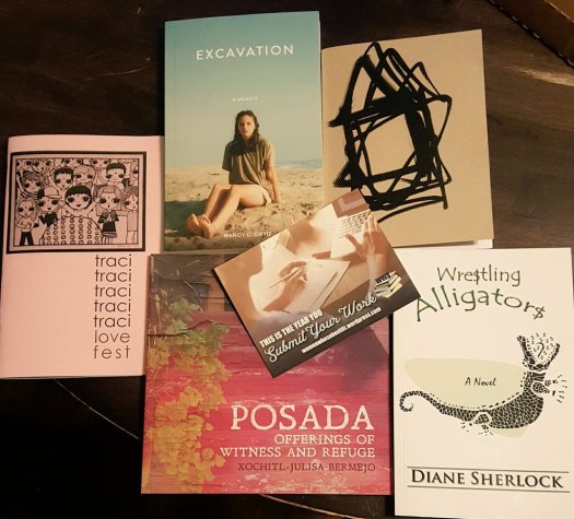 L-R in clockwise order: a zine title Love Fest, Excavation, a book by Wendy C. Ortiz, a chapbook with a beige cover with a black design, Wrestling Alligators, a book by Diane Sherlock, and Posada, a book by Xochitl-Julisa Bermejo