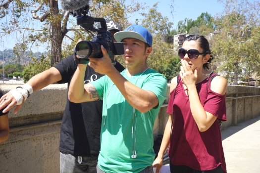 A man holds a video camera, a woman with sunglasses stands behind him, along with another man pointing, whose face is obscured