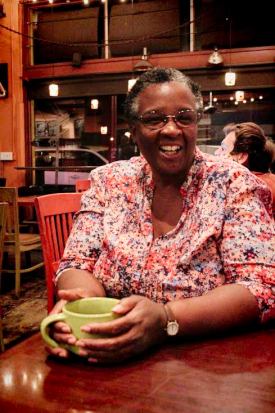 Half-shot photo of writer Desiree Kannel at a cafe, holding a green coffee mug, smiling