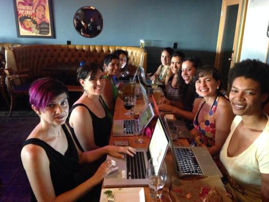 Eight women with laptops sit on either side of a long table, smiling at the camera