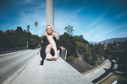 A white woman dressed in a black lace dress sits on a bridge railing, legs in front of her, holding a book; a tree-lined canyon and wash in the background